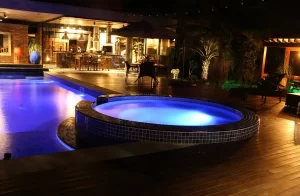 Piscina residencial iluminada à noite com luz LED azul, área de lazer moderna com deck de madeira, cadeiras e espaço gourmet ao fundo.