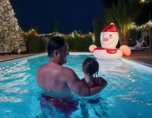 Pai e filha aproveitando brincadeiras de Natal na piscina à noite, com iluminação decorativa e boia natalina ao fundo