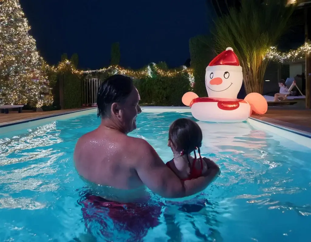 Pai e filha aproveitando brincadeiras de Natal na piscina à noite, com iluminação decorativa e boia natalina ao fundo